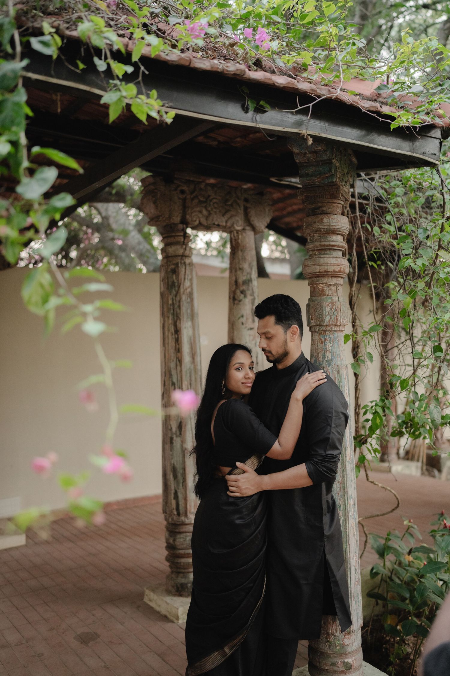 Couple in matching black Indian attire embracing under a stone pergola with lush greenery and pink flowers.