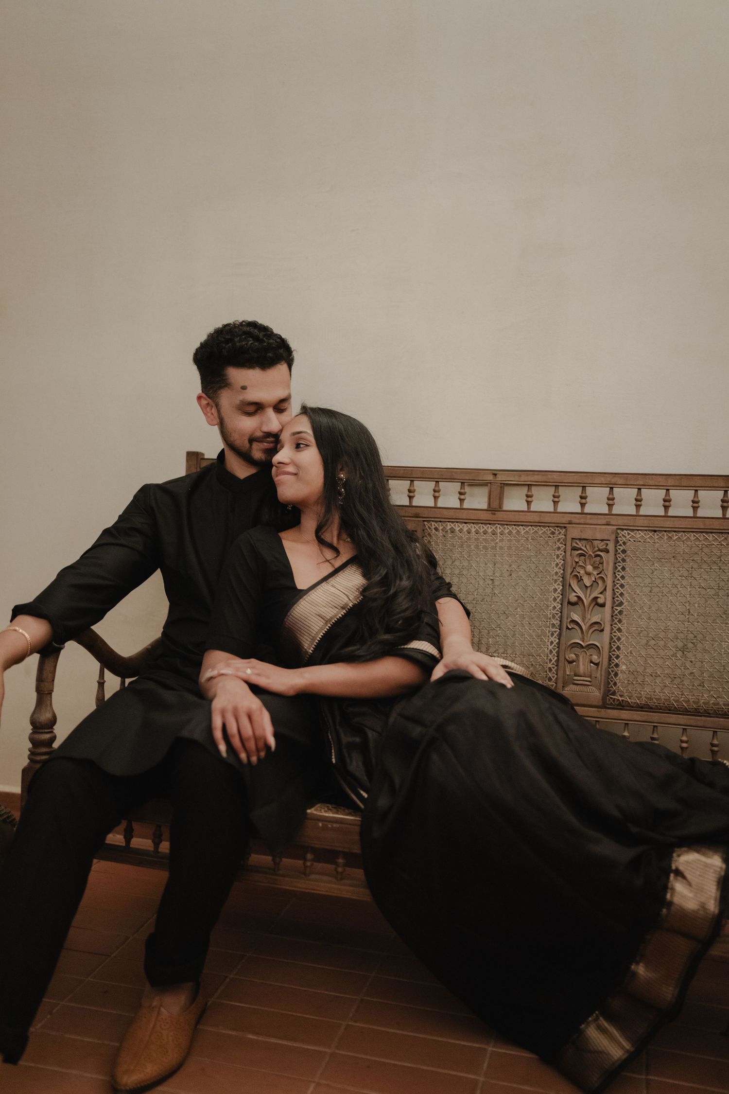 Couple in matching black outfits sitting intimately on a carved wooden bench, woman in saree leaning toward man.