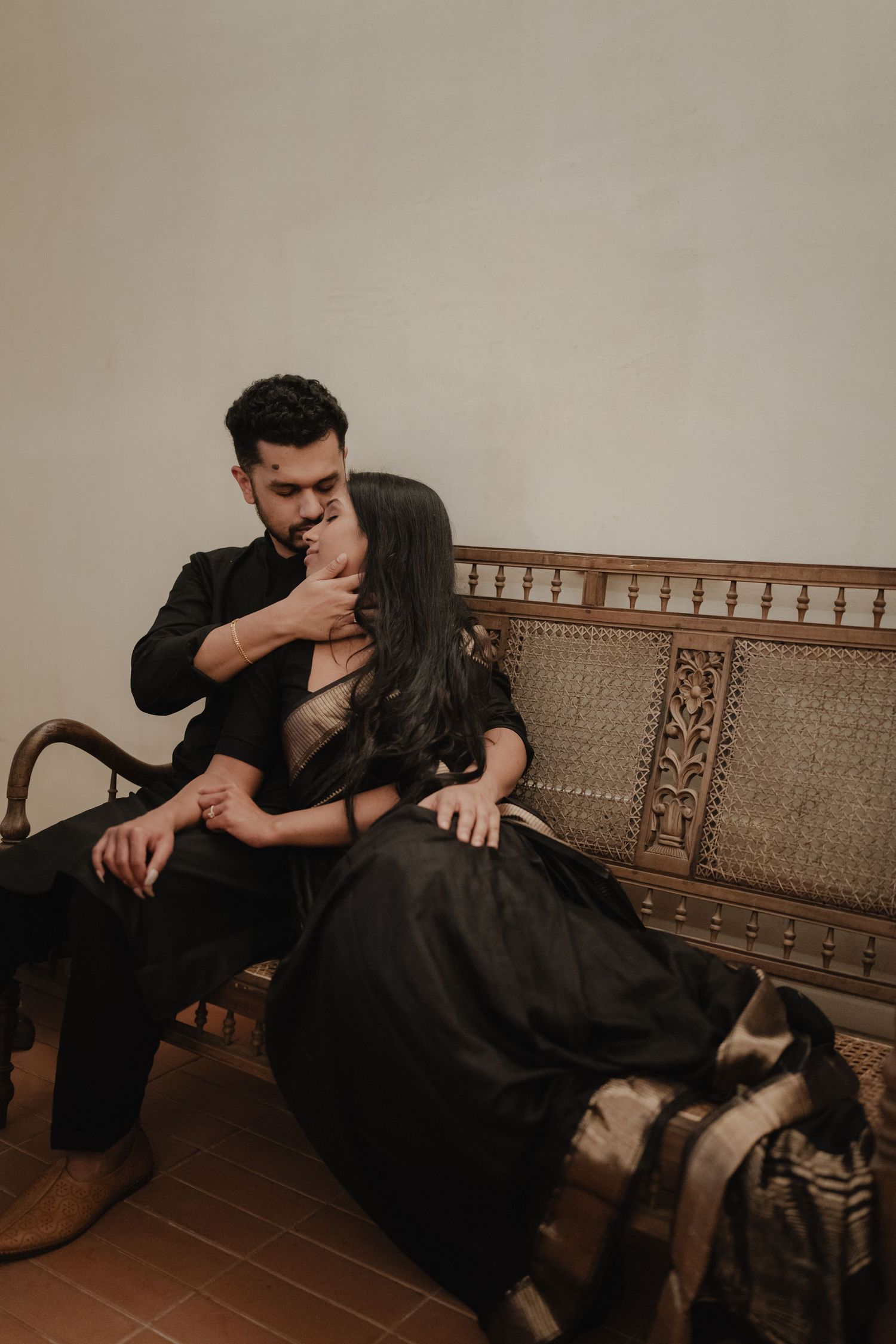 Romantic couple on ornate wooden bench, man in black kurta gently kissing woman's hand in elegant black saree.