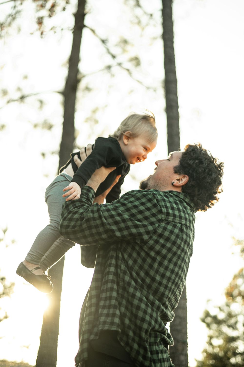 A playful outdoor moment in a forest backlit by golden sunlight as a child is lifted high into the air.