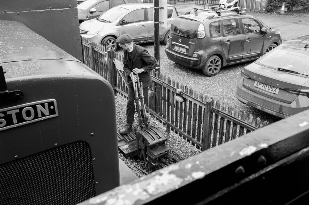 Black and white urban street scene showing parked cars and a metal fence with Bridgestone signage visible.
