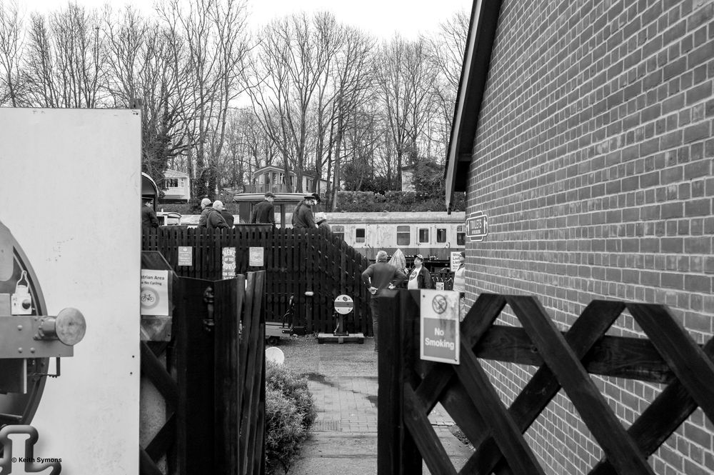 A black and white image shows a long brick wall with warning signs and trees visible in the background.