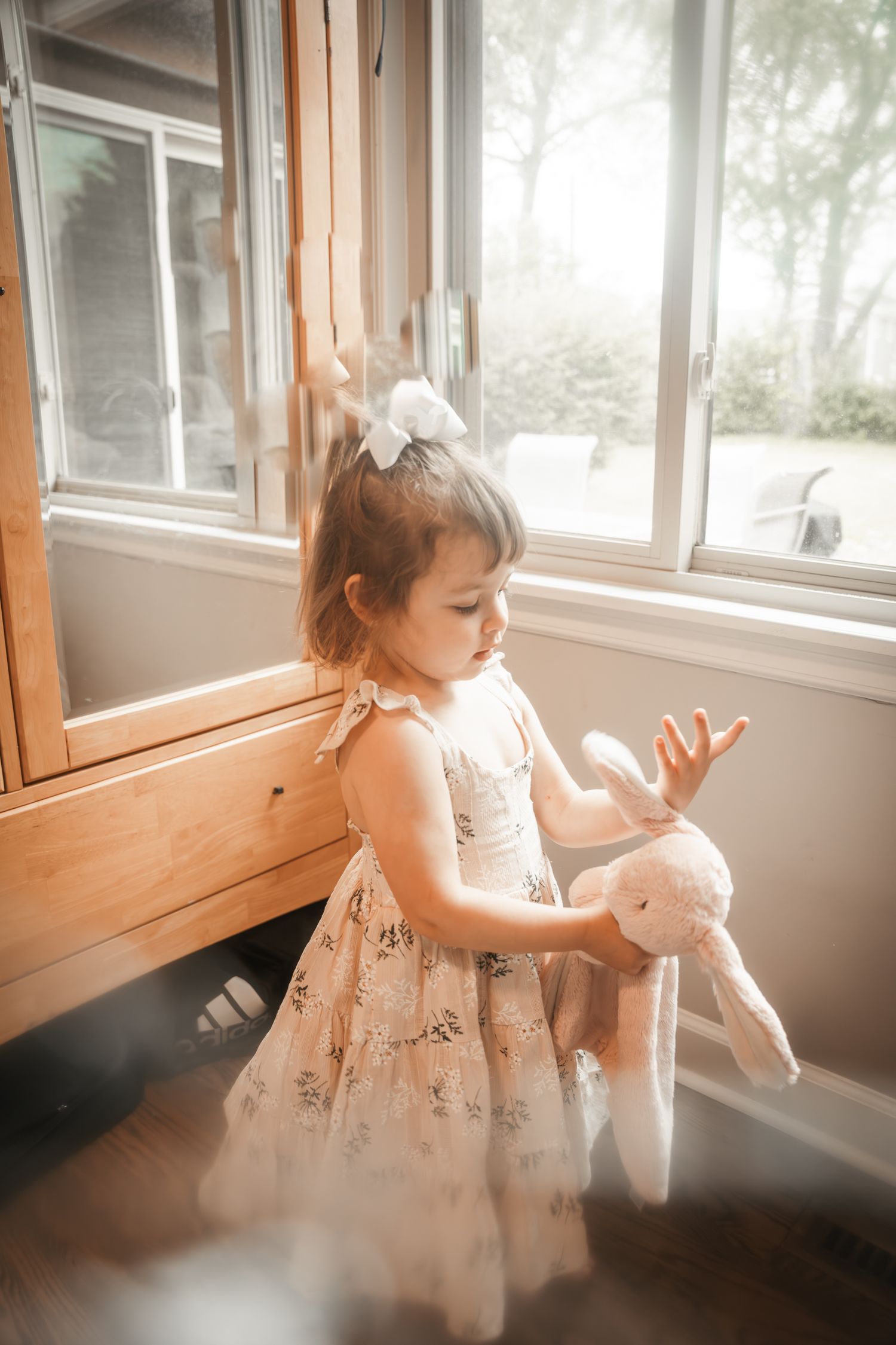 A small child in a floral dress plays with a plush bunny toy near a sunlit window in a dreamy, warm-toned setting during a film session
