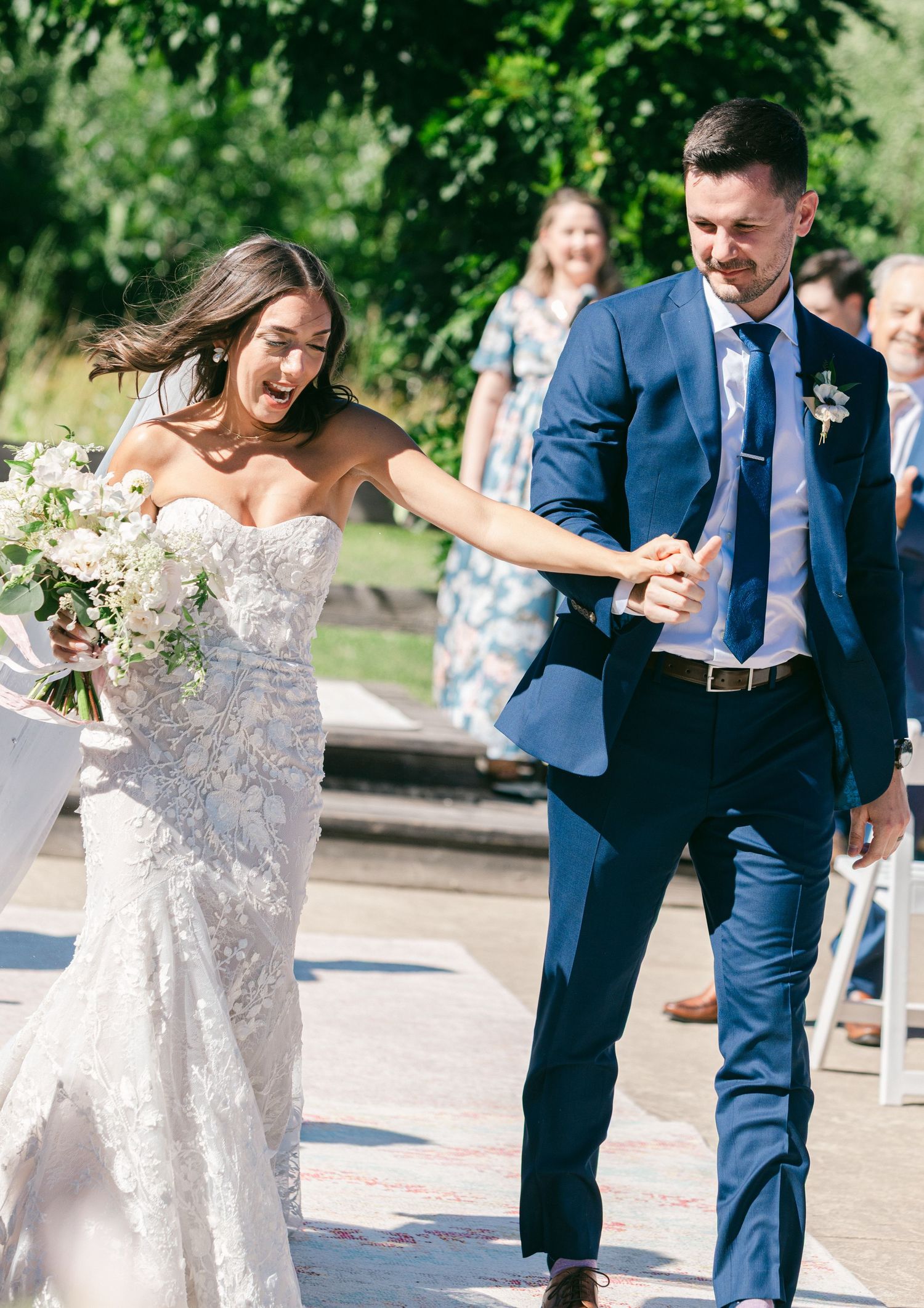 A couple in wedding attire dances together outdoors in a garden setting on a sunny day.