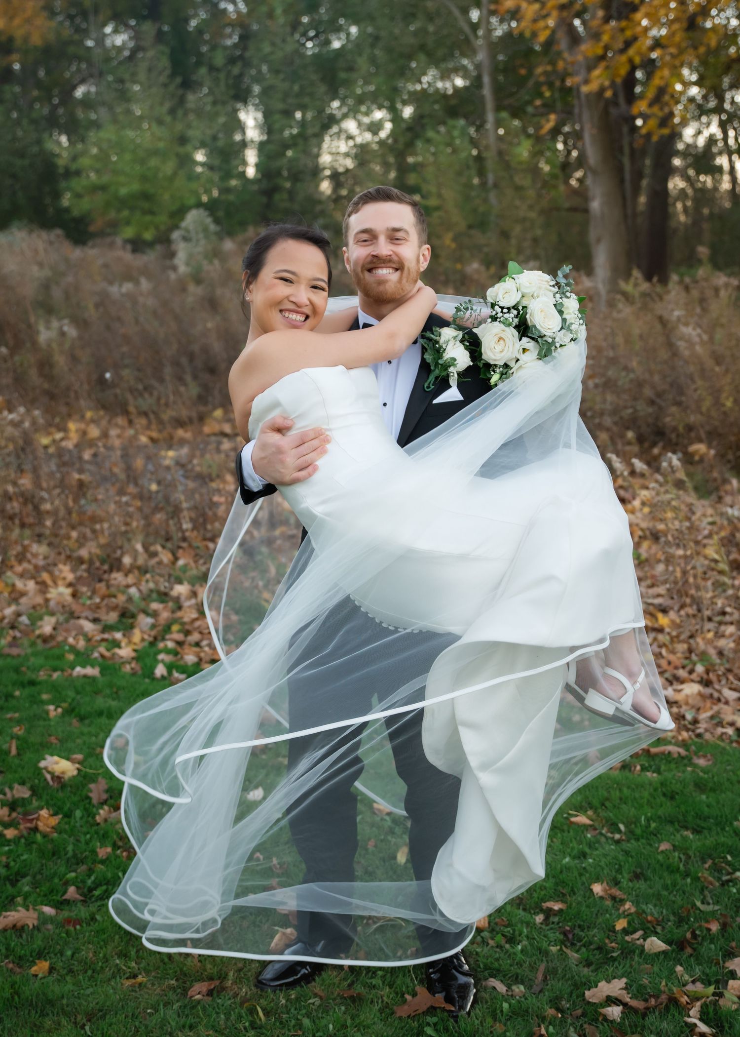 A wedding couple playfully dances together in a fall setting with autumn leaves scattered on the ground.
