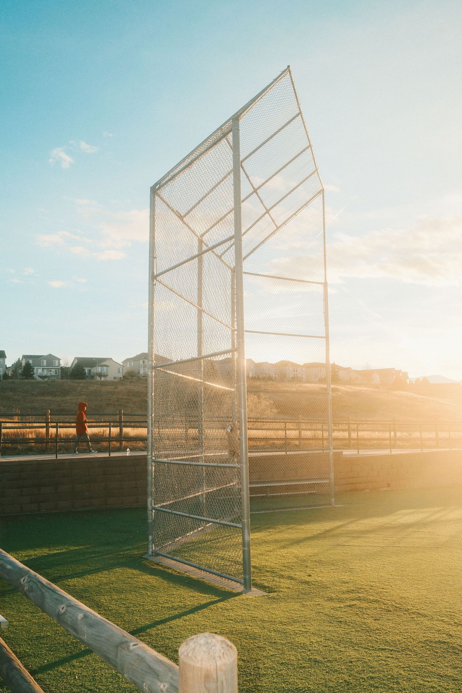 Baseball field backstop netting silhouetted against a golden sunset sky with green grass in the foreground.