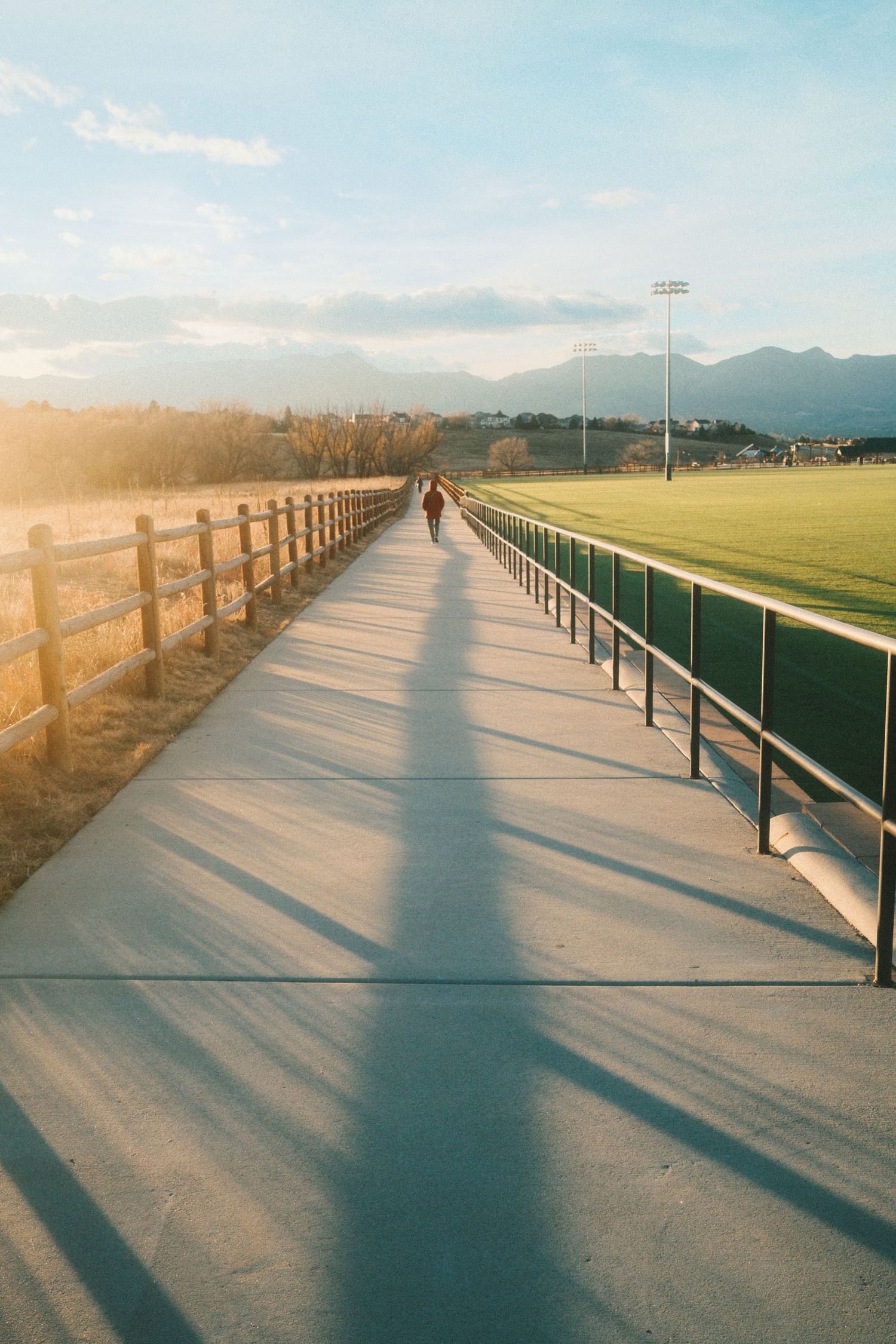 A person walks along a wooden fence-lined path at sunset with mountains and sports fields visible in the distance.