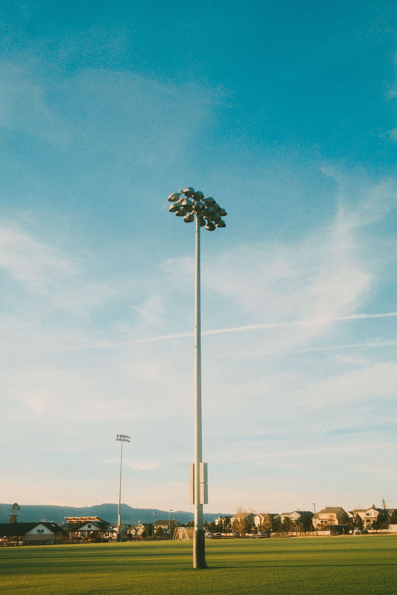 Tall light poles stand against a vibrant turquoise sky over an empty green sports field with houses in the background.