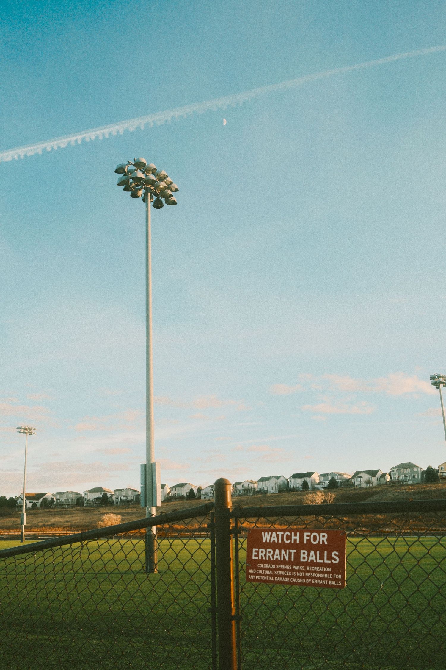 Warning signs for errant balls line a fence at a baseball field with suburban houses visible in the background.