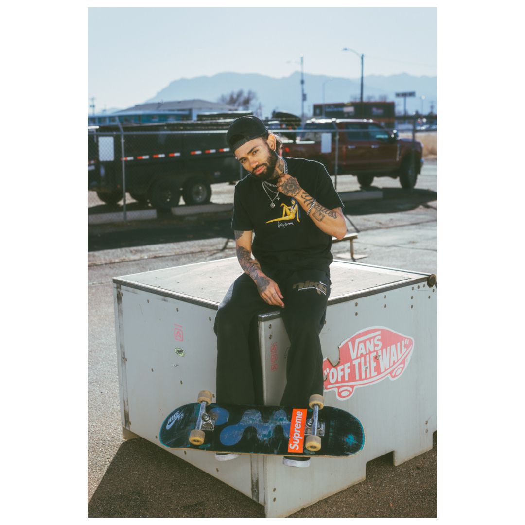 Tattooed skateboarder sitting on a Vans branded ramp with a skateboard, wearing a black beanie and t-shirt outdoors.