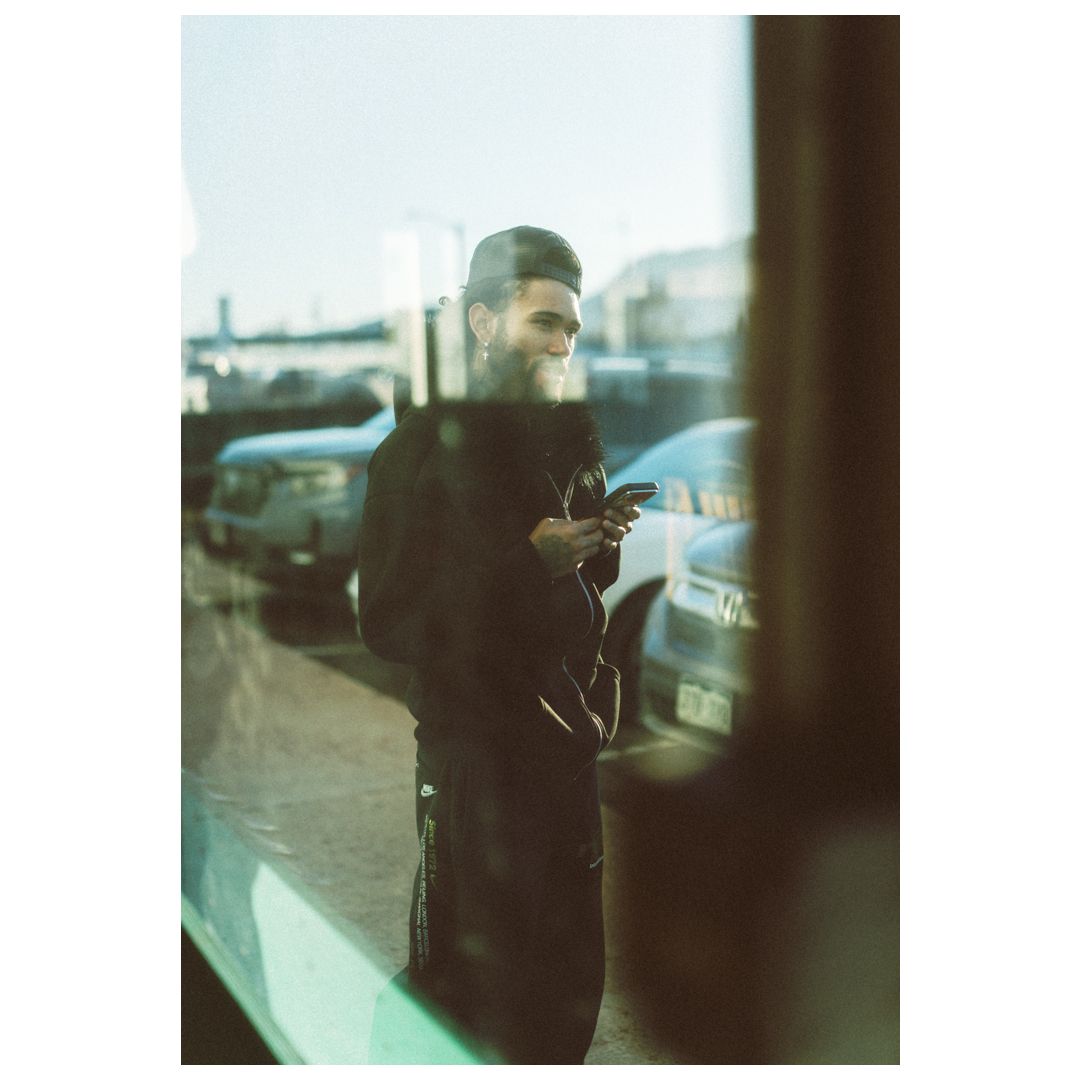 Bearded man in black coat using smartphone outdoors near parked cars on a sunny day.