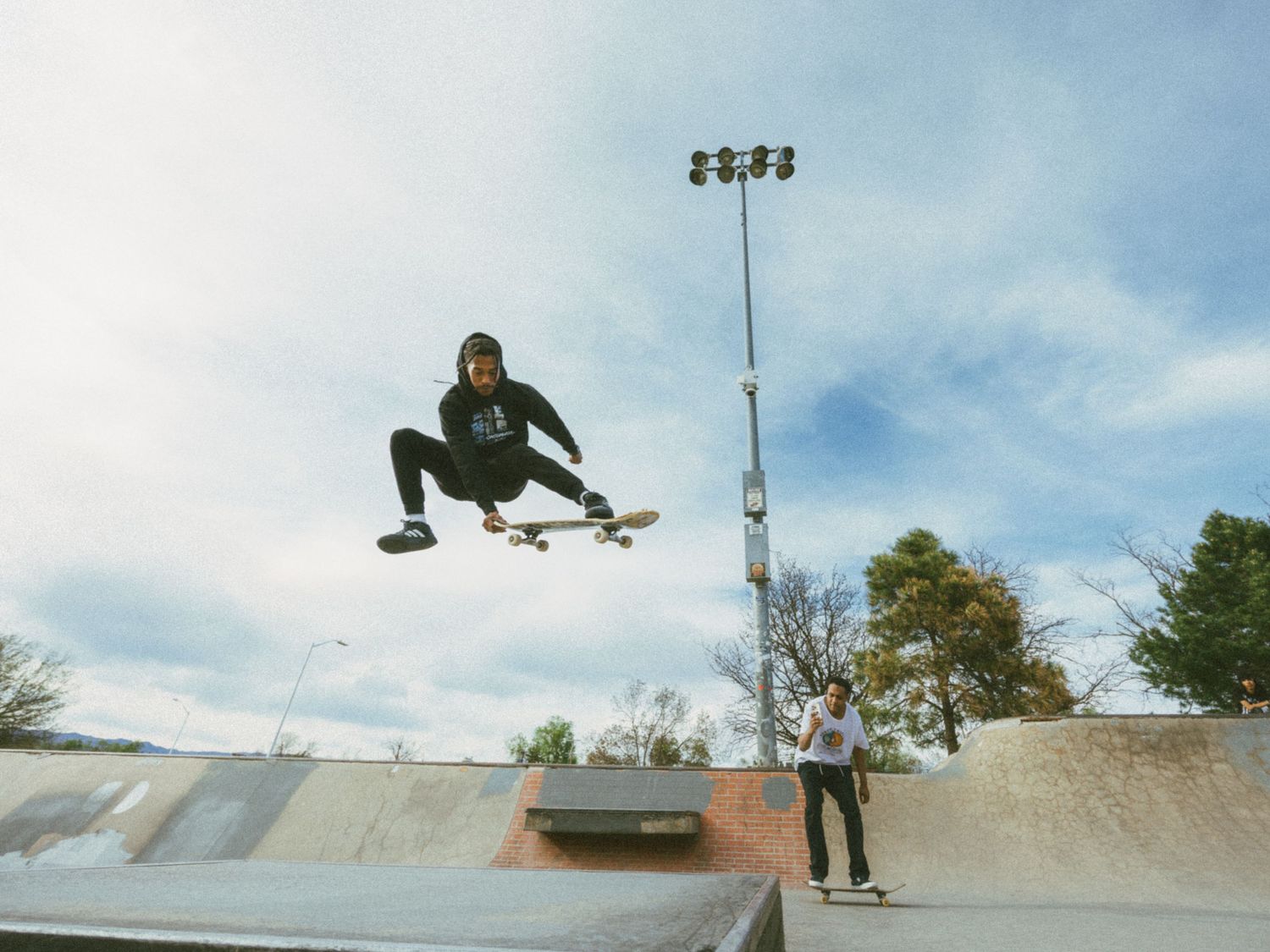 Skateboarder performing a high aerial trick at an outdoor skate park ramp under a partly cloudy sky.