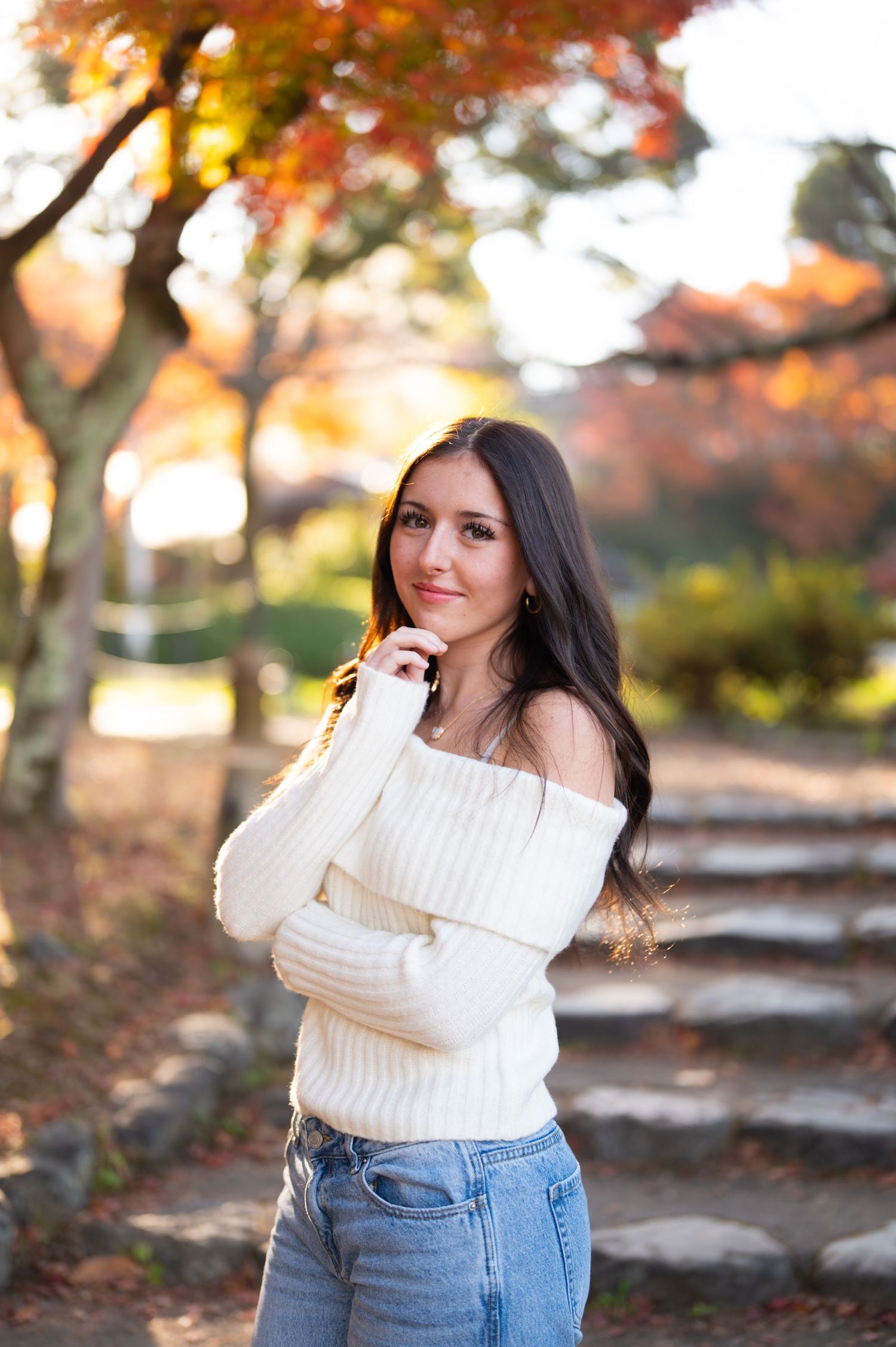 Young woman in off-shoulder white sweater posing with autumn foliage in the background.