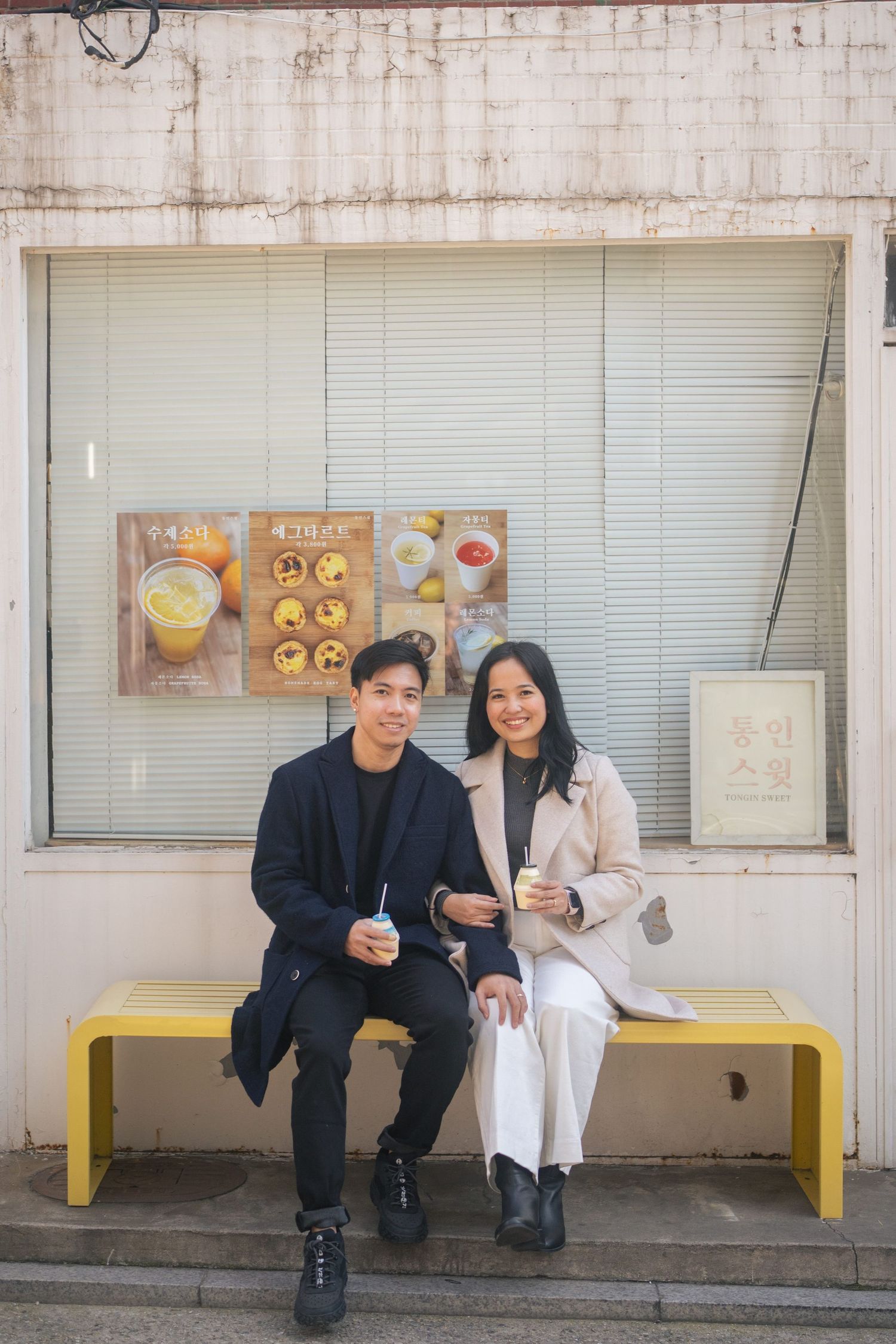 Two people sit on a yellow bench outside a storefront sharing drinks and enjoying a casual moment together.