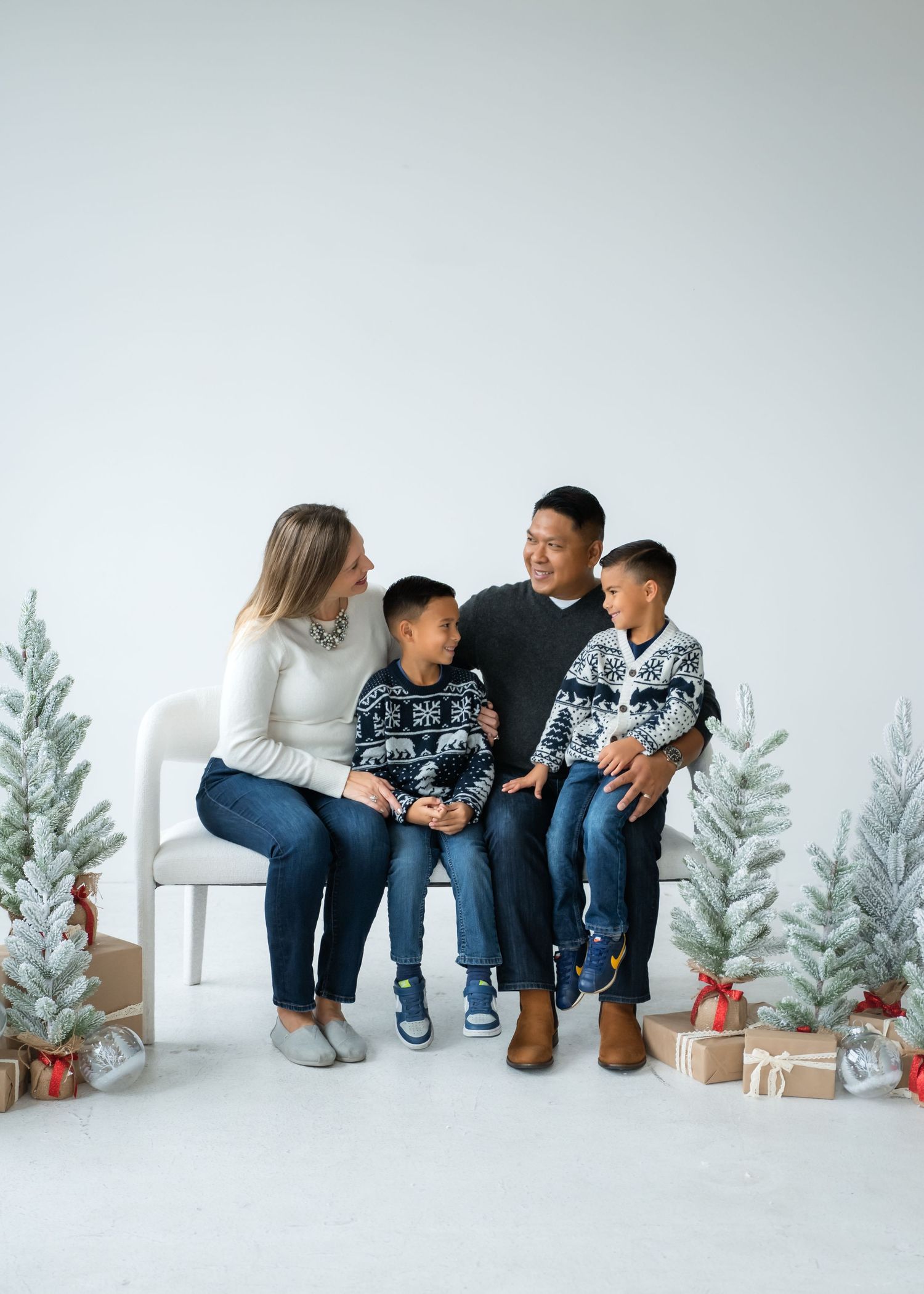 Family wearing matching winter sweaters sits together on white bench with holiday decorations and gifts around them.
