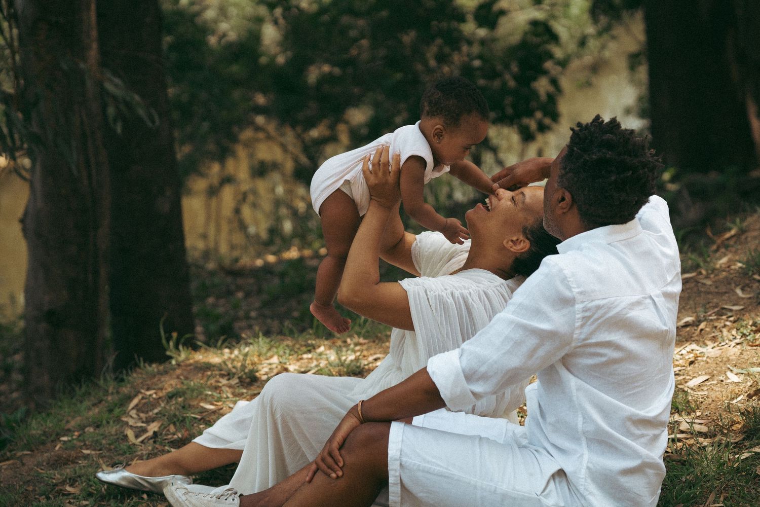 A couple in white clothing sharing playful moments in a sun-dappled forest setting.