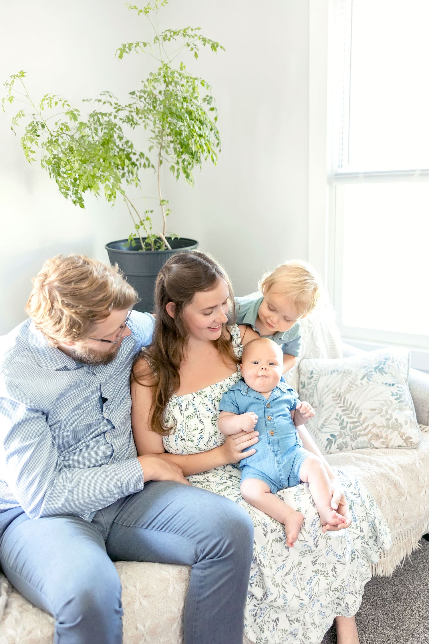 A family sits together on a couch in coordinated blue and white outfits while sharing tender moments admiring their new baby brother.