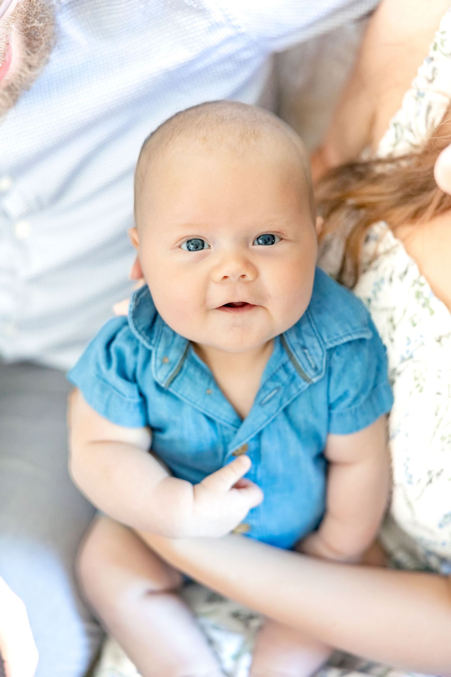 A baby wearing a blue shirt lies on white bedding while looking up with bright eyes and a gentle smile.