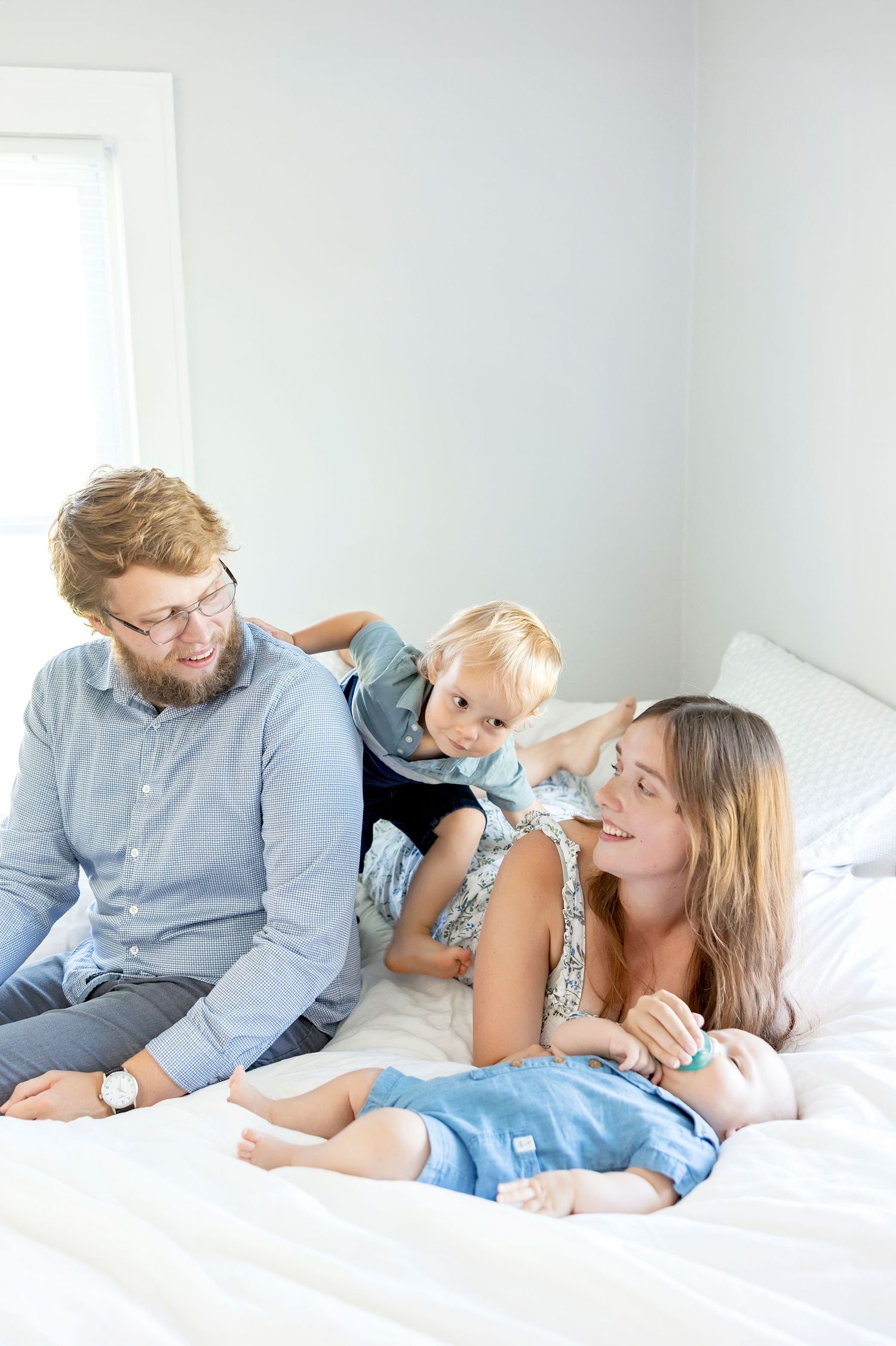 A family shares tender moments together while relaxing on a white bed in a bright room during Clear Lake in-home lifestyle session.