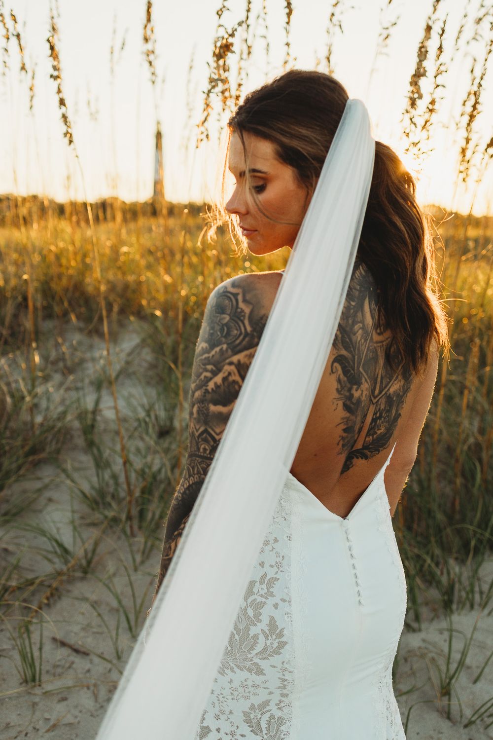 Bride in a lace wedding dress with flowing veil poses against sunset beach dunes.