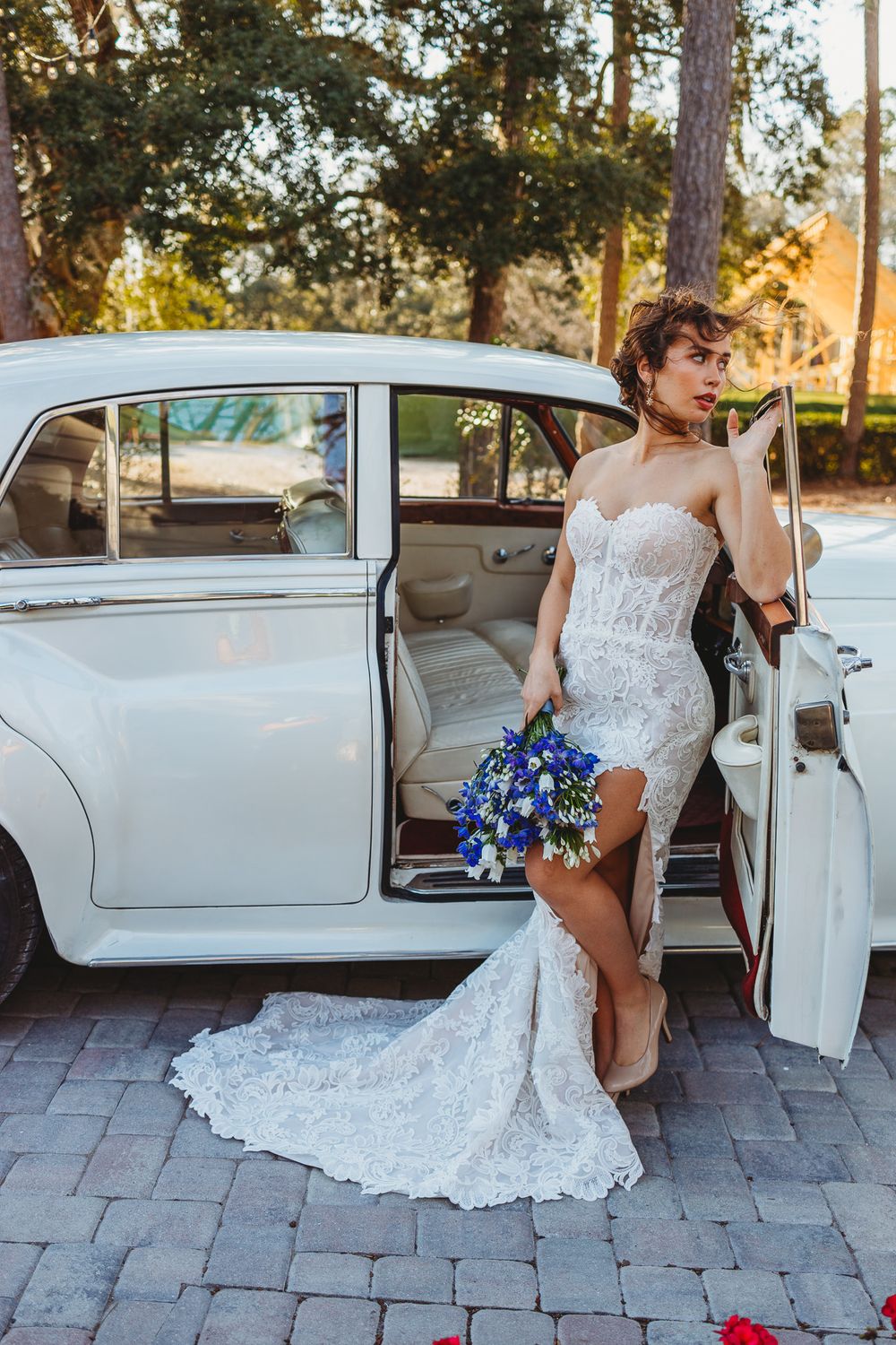 Elegant bride in lace mermaid wedding dress poses with vintage white car at sunset.
