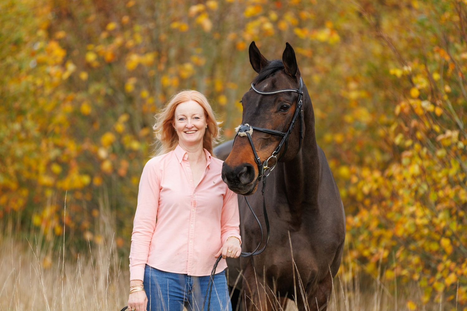 A person in a pink sweater and jeans stands with a black horse against a backdrop of golden autumn foliage.