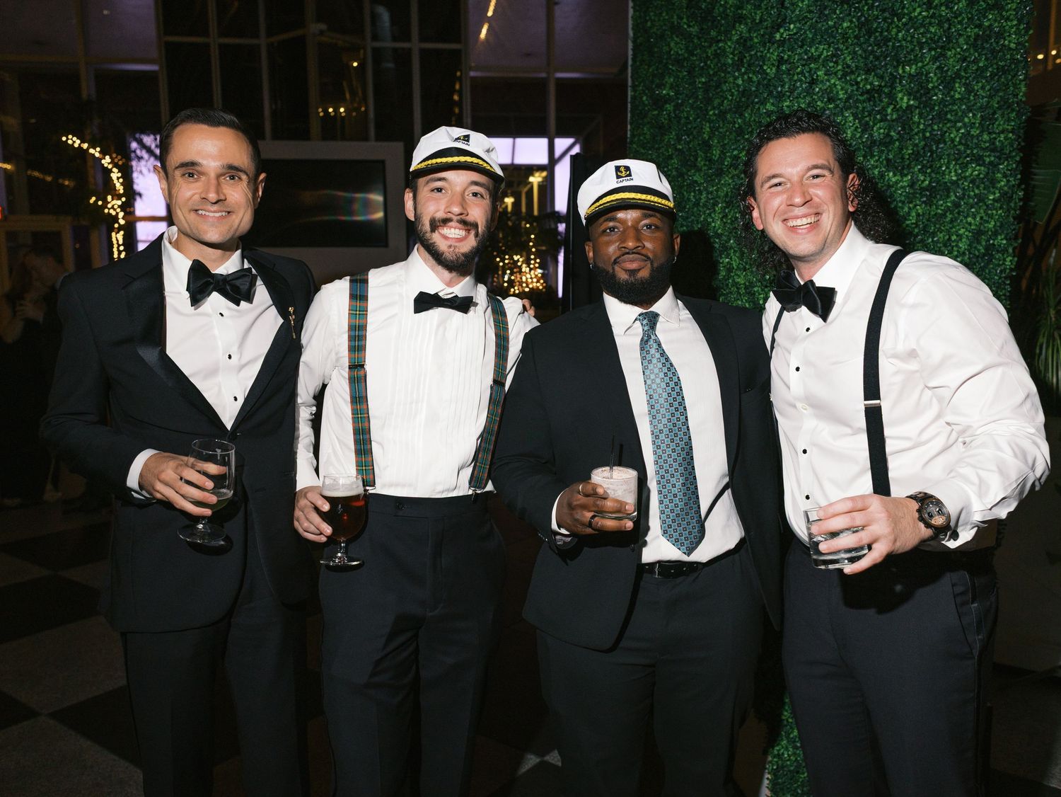 Four well-dressed men in formal attire with bowties and suspenders smile while holding drinks at an elegant event.