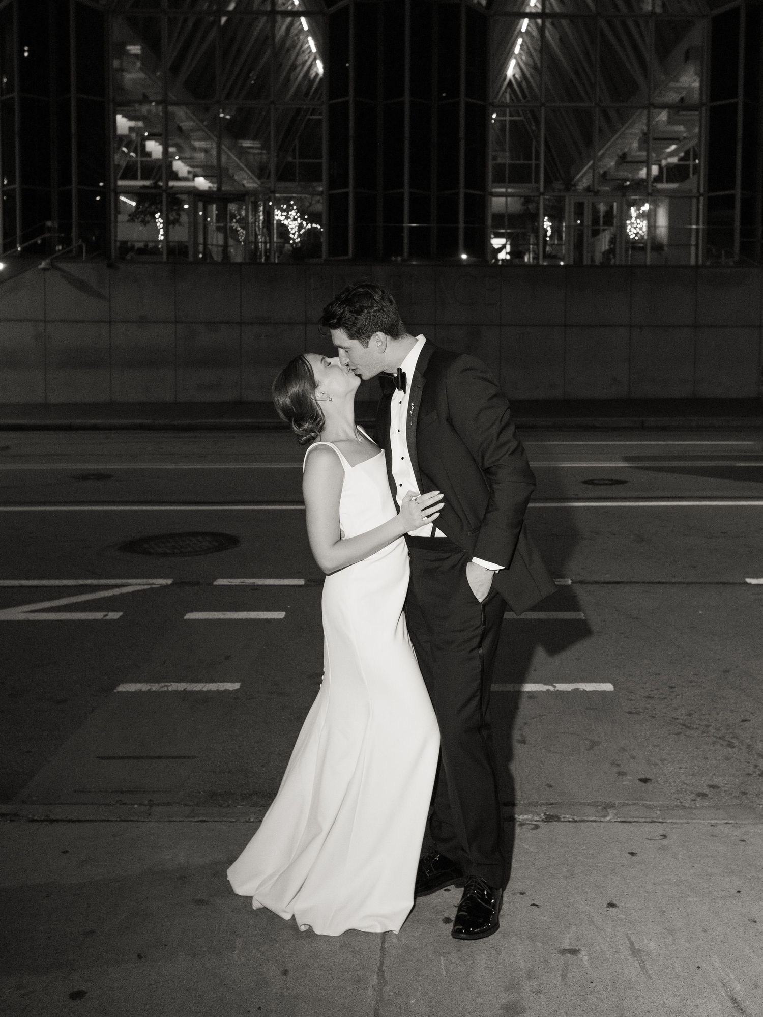 A couple in formal attire dances together on a city street at night, creating a romantic black and white scene.