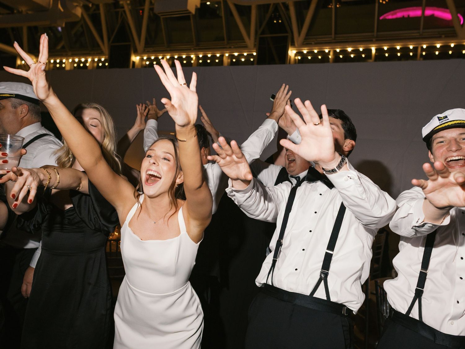 Wedding party guests dance and celebrate with raised arms on a dimly lit dance floor at a formal reception.