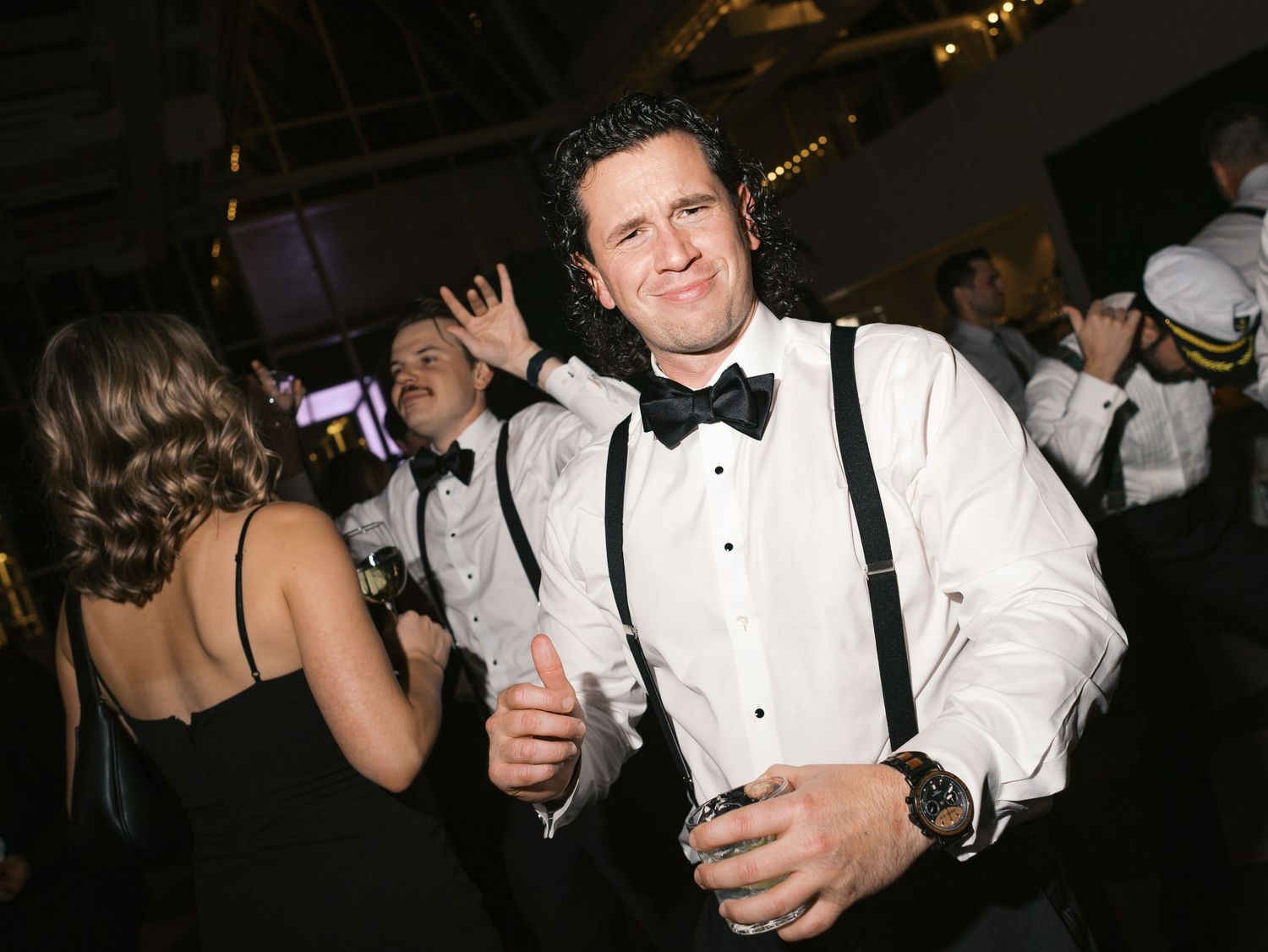 A person in a white tuxedo shirt and suspenders dances at a formal event with drinks in hand.