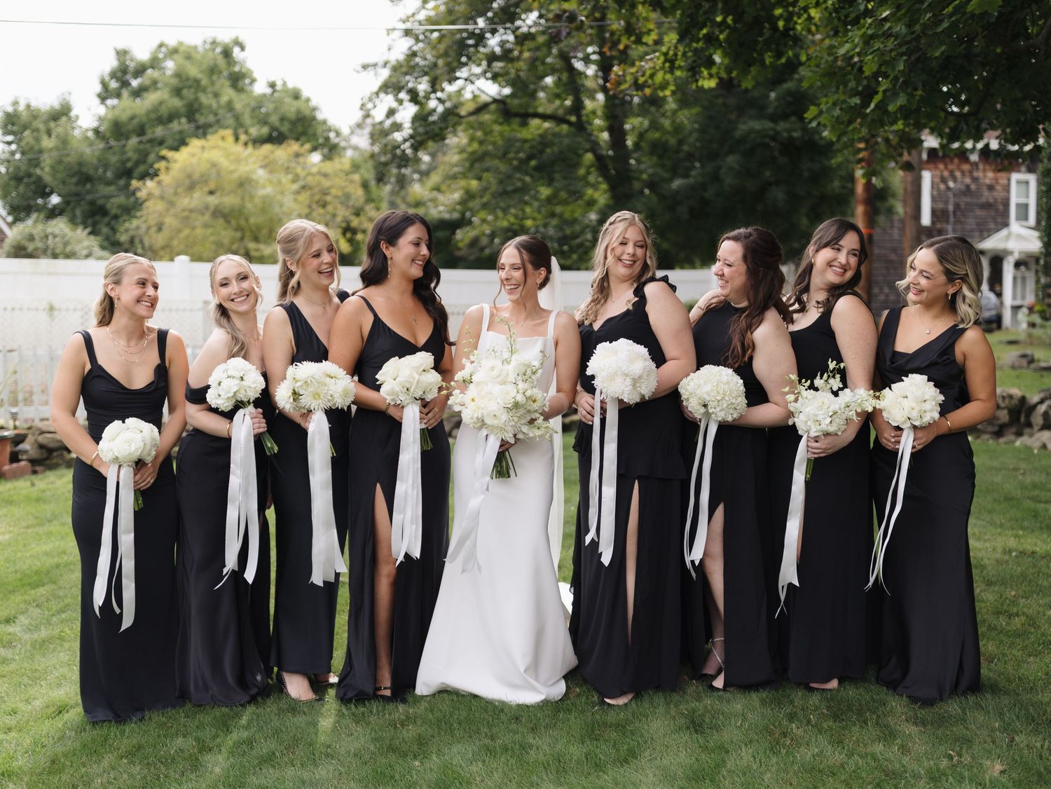 Wedding party in black bridesmaid dresses and white bouquets posing outdoors on a sunny day.