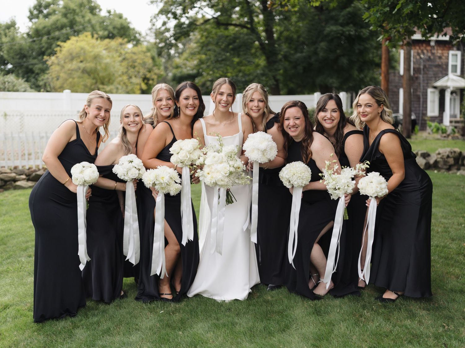 Bridal party in black dresses lined up on green lawn holding white floral bouquets.