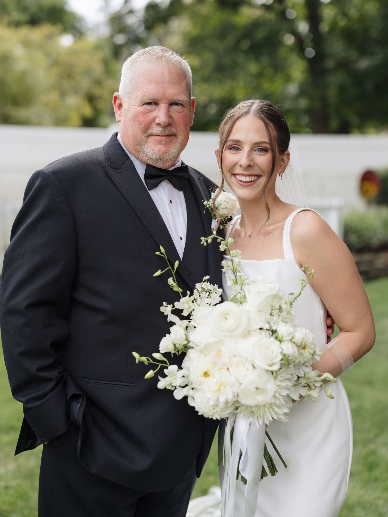 A wedding couple poses with family members outdoors in formal attire holding white floral bouquets.