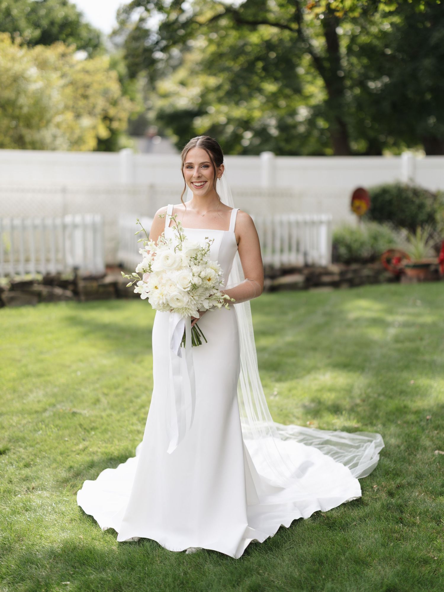 Bride in flowing white wedding gown holding white bouquet on sunny garden lawn.