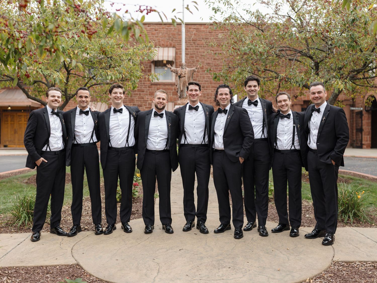 Group wearing black tuxedos and bowties posed together outdoors at formal event.