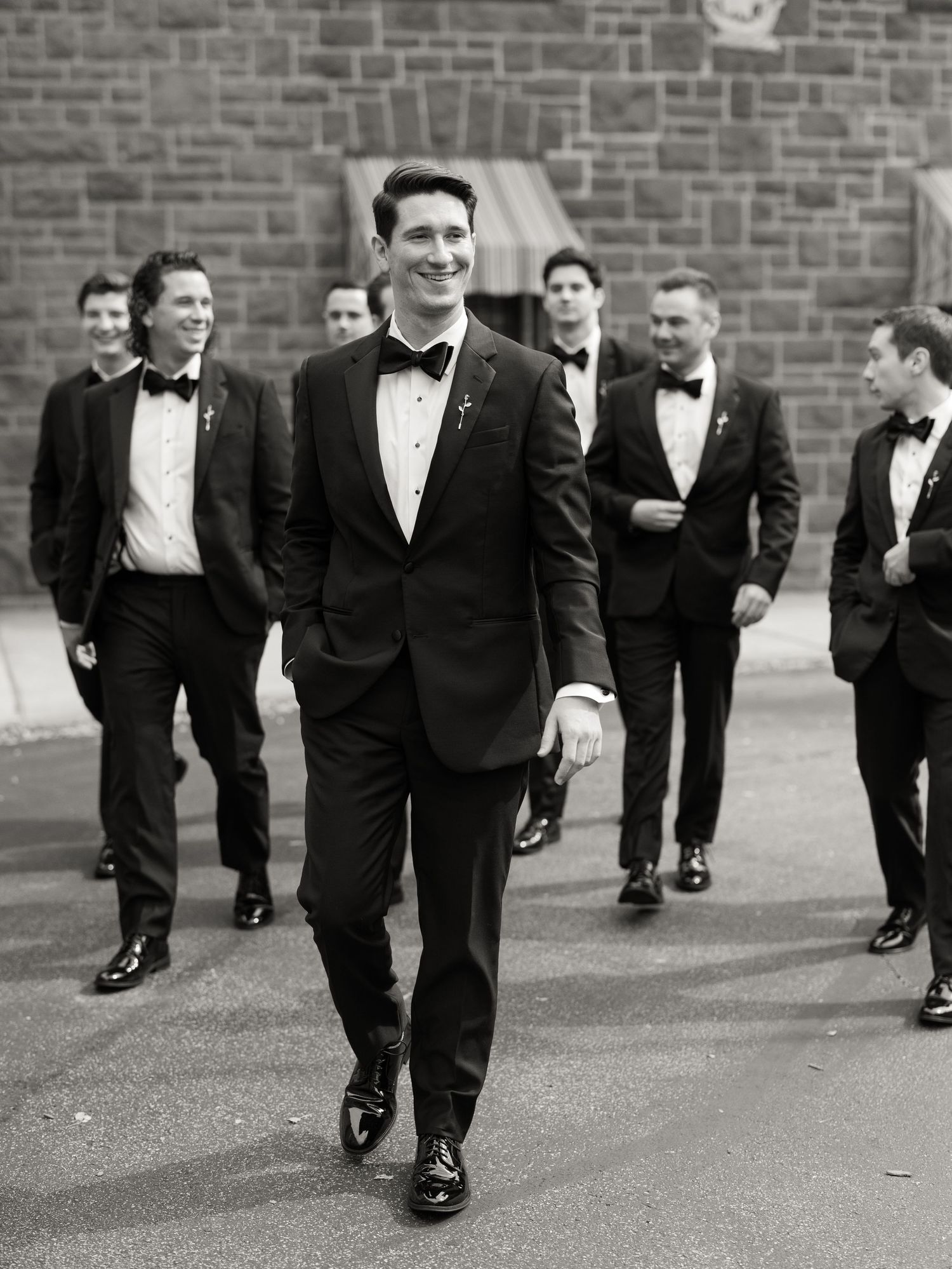Black and white photo of groomsmen walking in a line wearing tuxedos against a brick wall background.