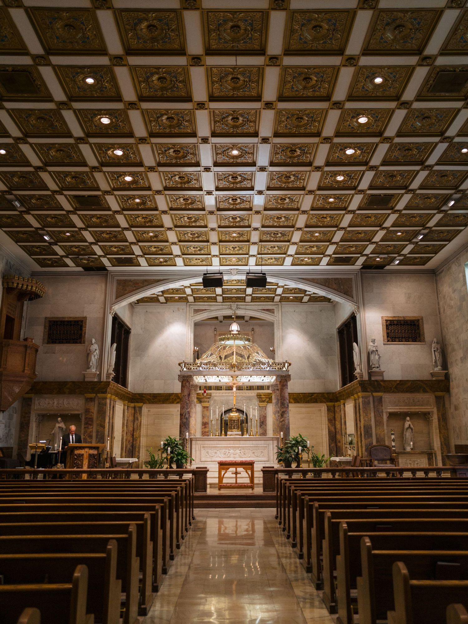 Elegant church interior with ornate coffered ceiling, wooden pews, and gilded altar beneath an arched sanctuary.
