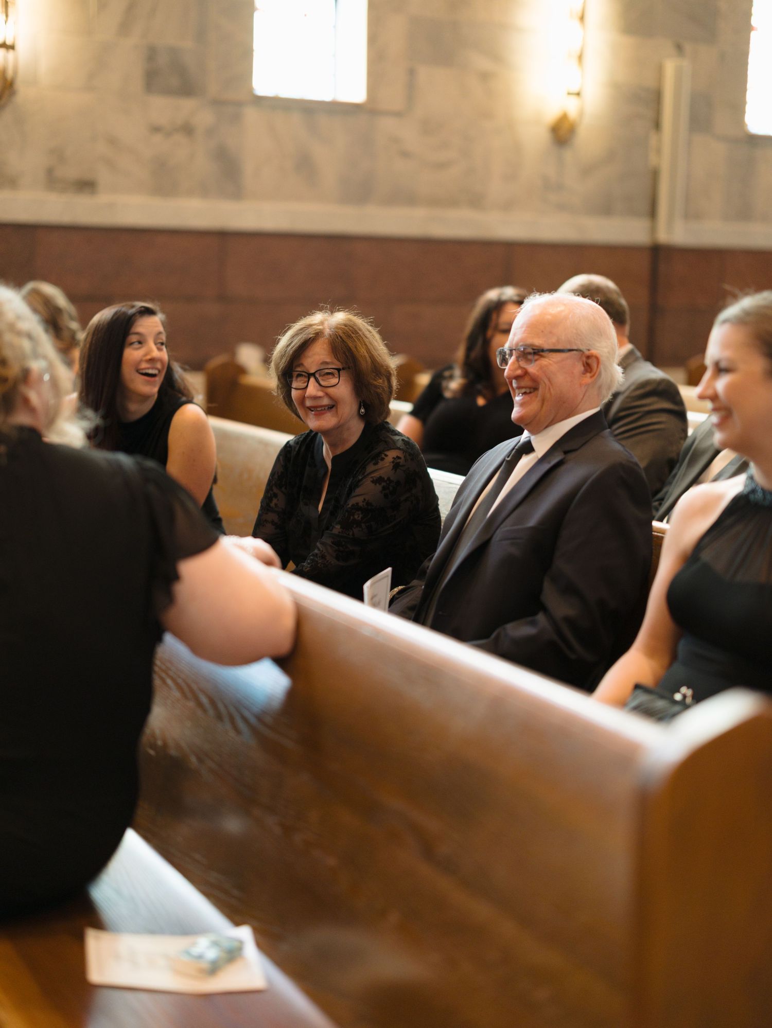 Wedding guests seated in wooden church pews smile and laugh during a ceremony.