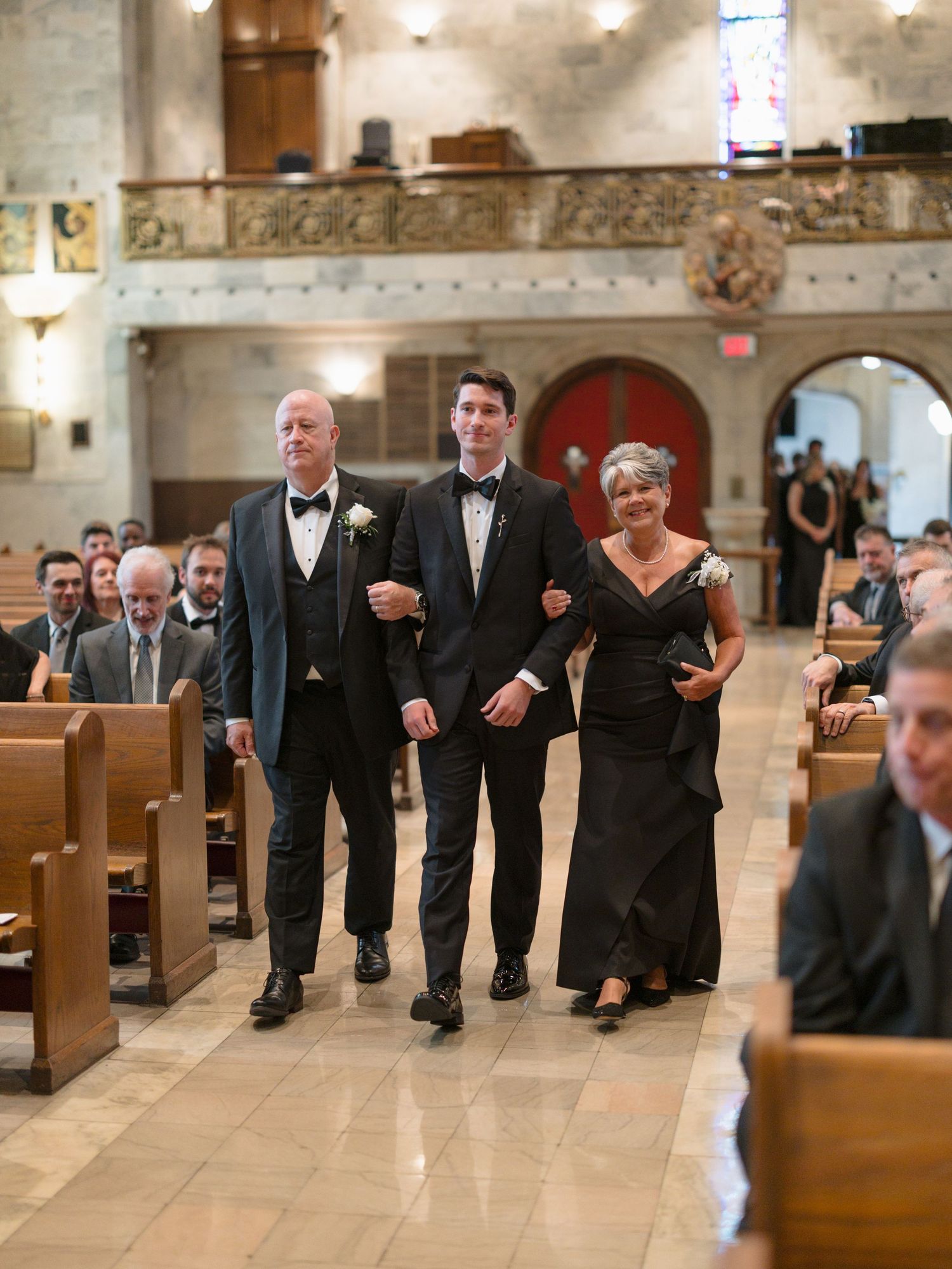 Groomsmen in black tuxedos walk down church aisle in playful synchronized formation during wedding ceremony.