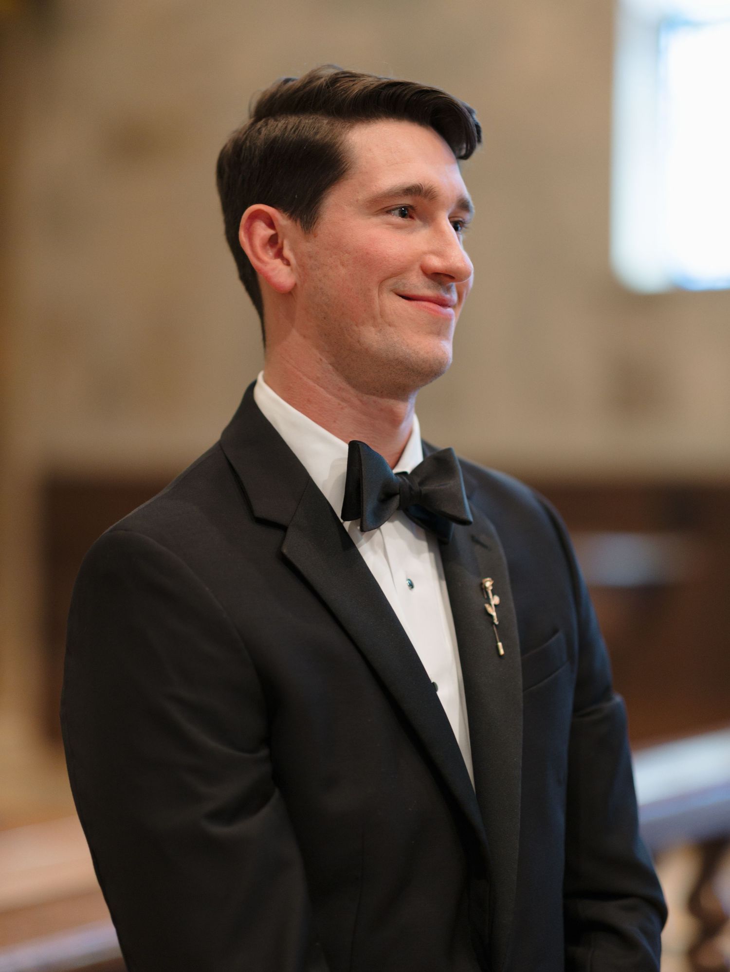 Groom in classic black tuxedo and white bow tie smiles during wedding ceremony.