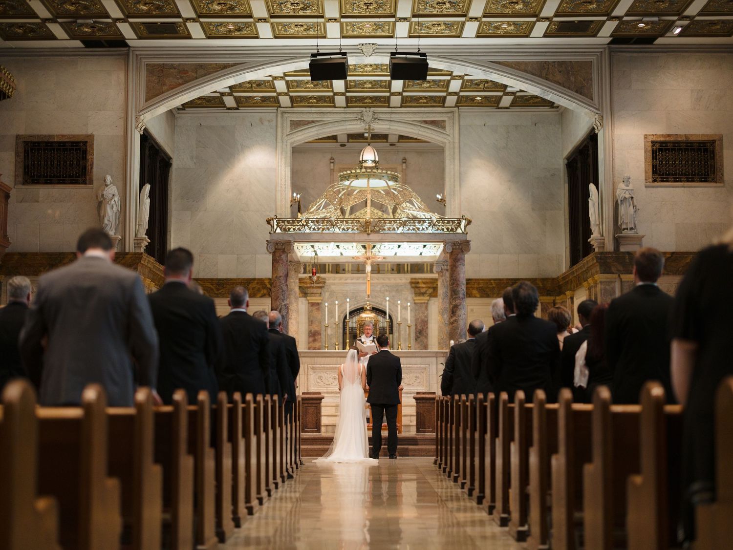 Elegant church wedding ceremony with guests seated in wooden pews under an ornate arched ceiling with chandeliers.