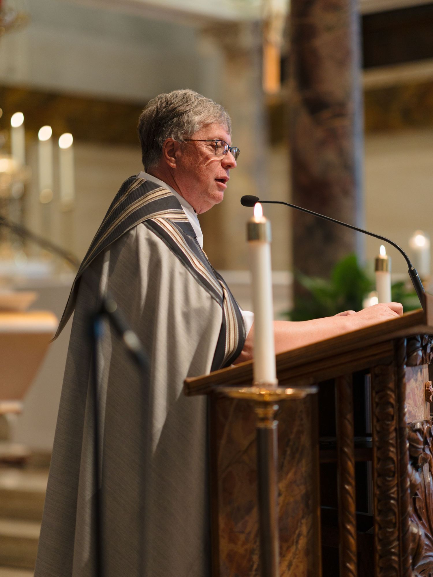 A priest in gray robes stands at a wooden church podium during a ceremony.