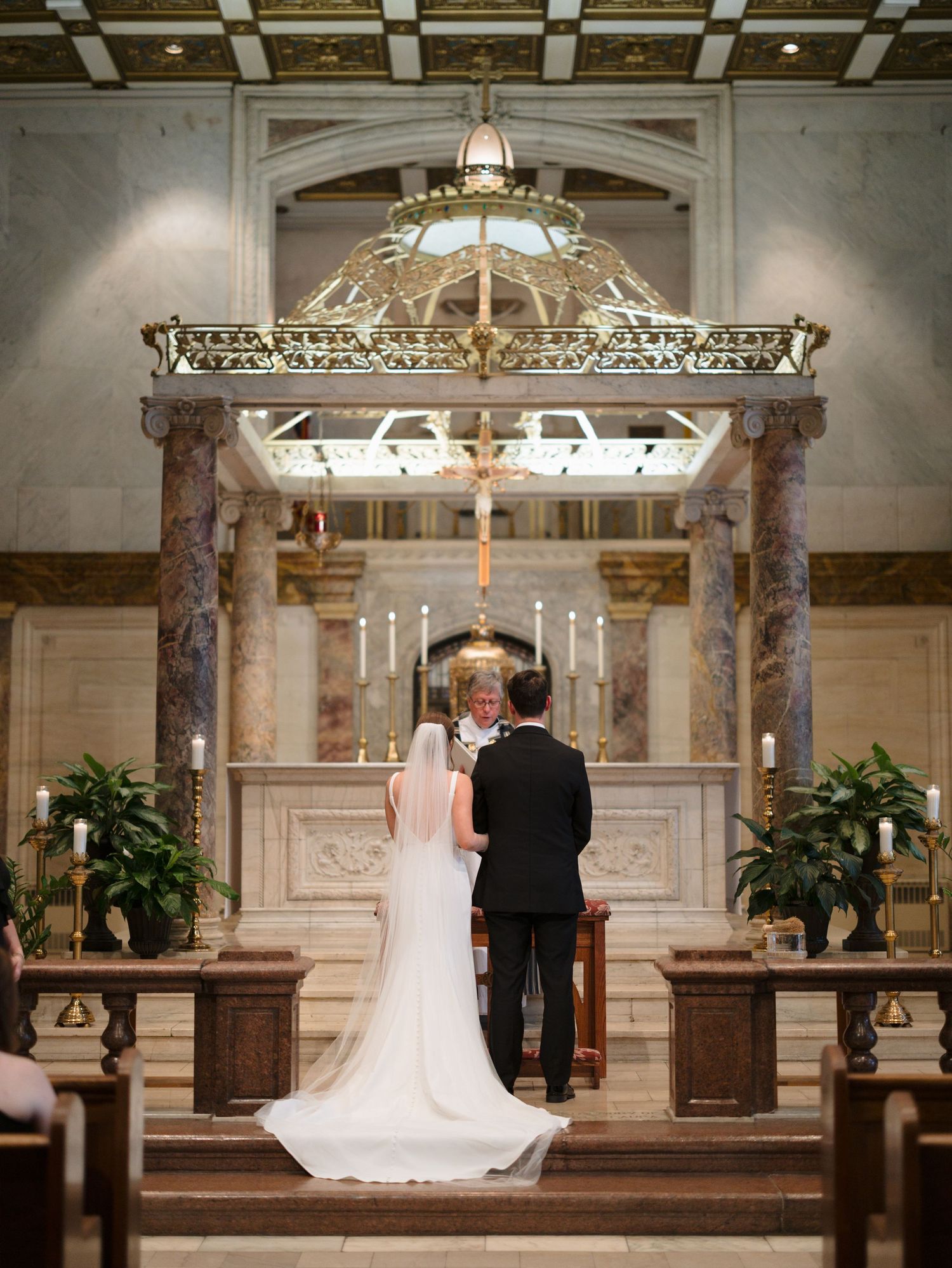 A wedding ceremony takes place under an ornate altar with marble columns and chandelier lighting.