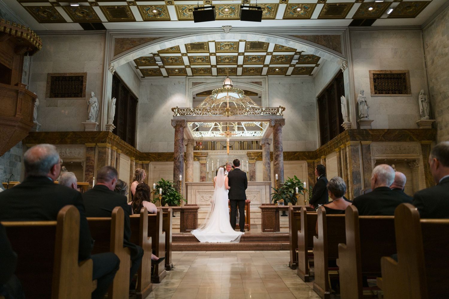Elegant wedding ceremony in a grand church with ornate coffered ceiling and chandeliers.
