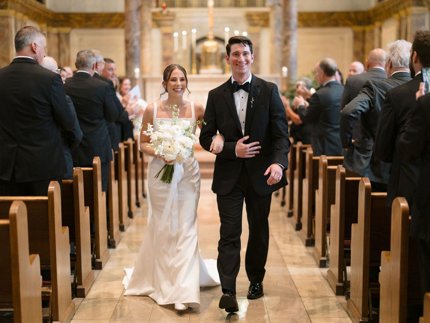 Wedding party members walk down the center aisle of a grand church with wooden pews on both sides.