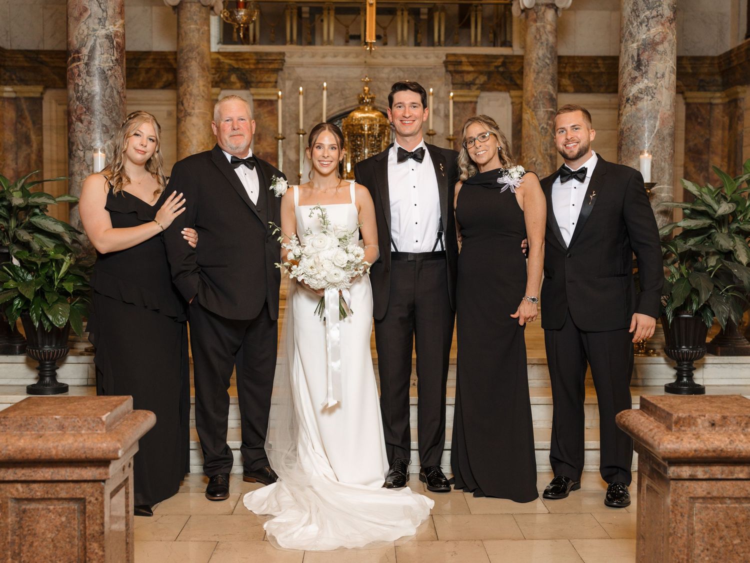 Wedding party in formal black attire and white gown posed in elegant church setting.