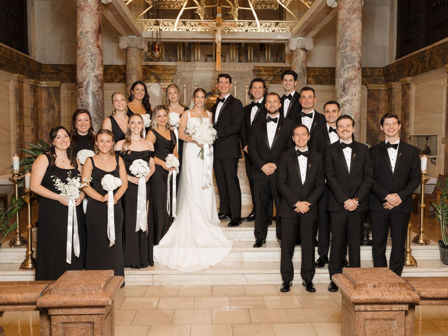 Wedding party in black and white formal attire posed on church steps with ornate altar backdrop.