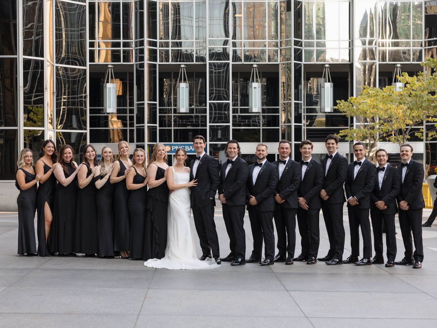 Large wedding party poses in front of modern glass building with bridesmaids in black and groomsmen in tuxedos.