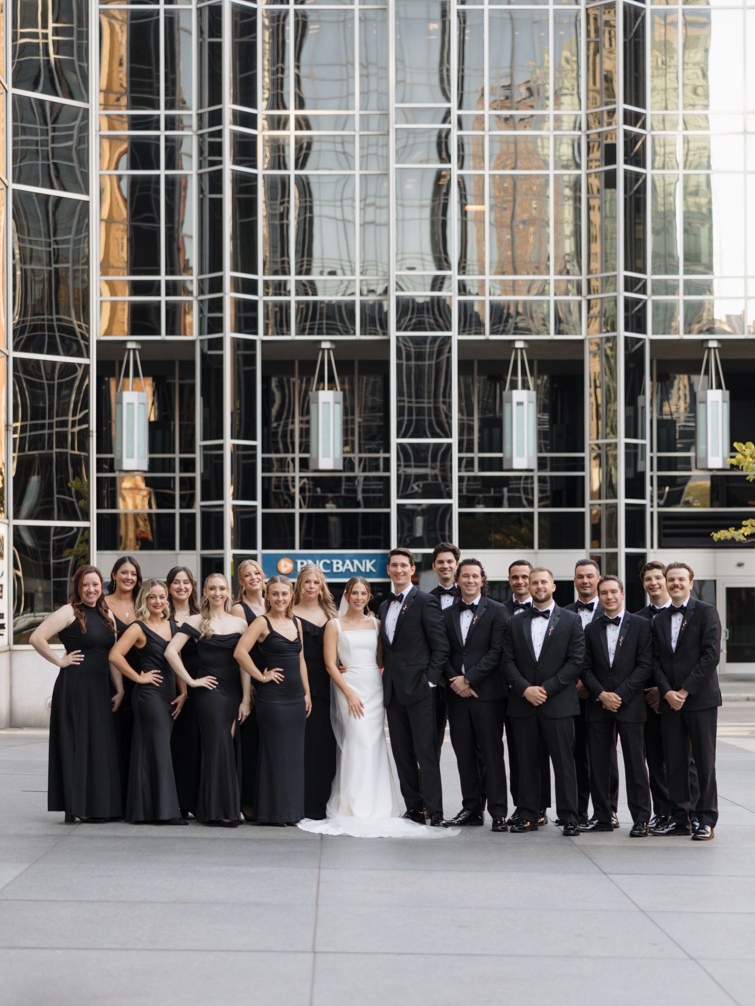 Large wedding party in formal black attire poses in front of modern glass building.