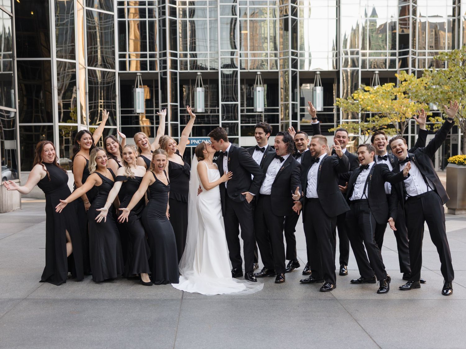 Wedding party in formal attire poses together outdoors near modern glass building.